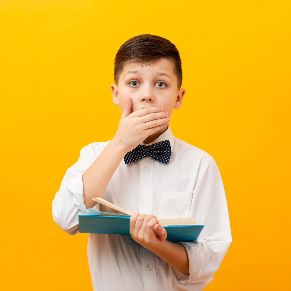 A child astonished by forgetfulness A young boy wearing a white dress shirt and a blue patterned bowtie stands against a solid bright yellow background. He holds an open blue book while covering his mouth with his right hand, looking directly ahead with wide, surprised eyes.