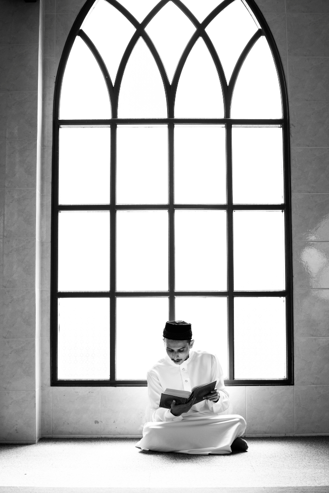 A black and white photograph of a young man sitting cross-legged on the floor, deeply focused on reading an open book held in his hands. He is positioned in front of a massive, arched multi-pane window that fills the background with bright, soft light.
