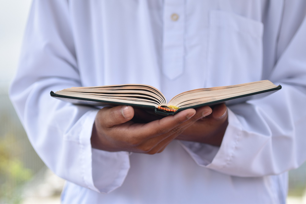 A close-up shot focusing on the hands of a person wearing a white traditional thobe, gently holding open a book with a dark cover. The person's face is out of frame, and the background is softly blurred.