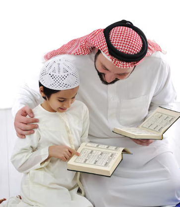 A child reviewing the Quran with his father A man in a white thobe and a red-and-white checkered ghutra sits closely with a young boy wearing a white thobe and a white kufi cap. Both hold open copies of the Quran, and the man smiles warmly while looking down as the boy points to a page.