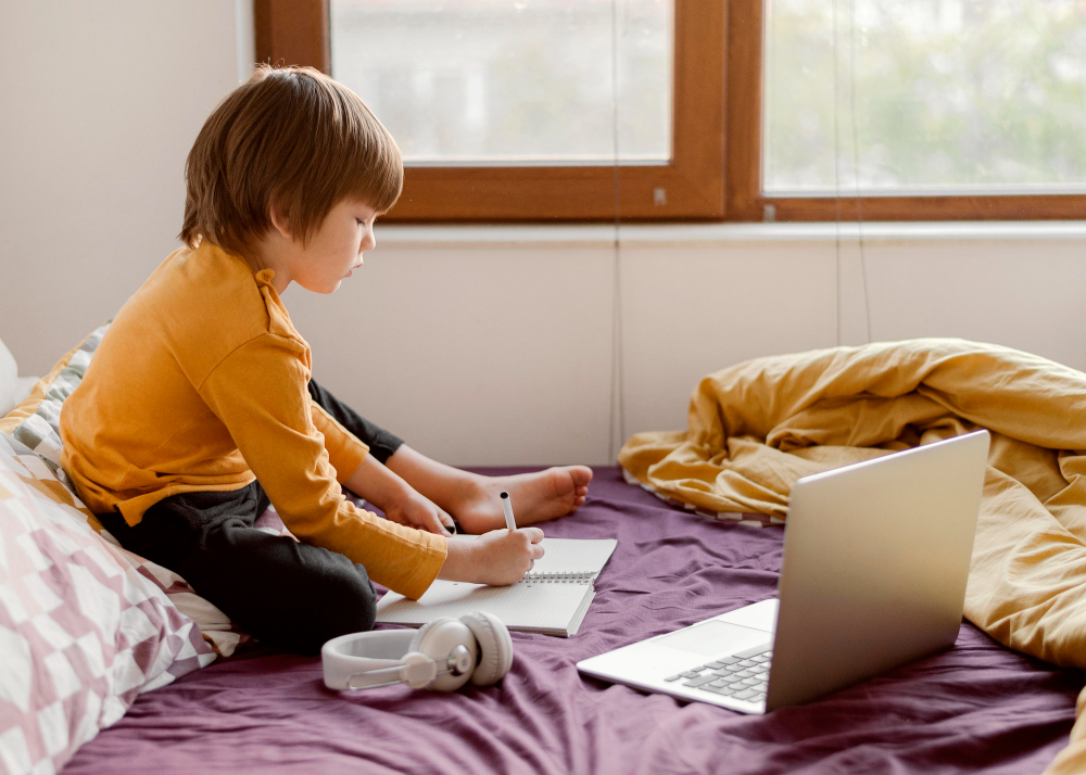 How Flexible Scheduling Heals the Home Environment for kids A young boy in a yellow long-sleeved shirt sits on a purple bed, focused on writing in a spiral notebook with a pen, while a laptop rests open in front of him and a pair of white headphones lies nearby.