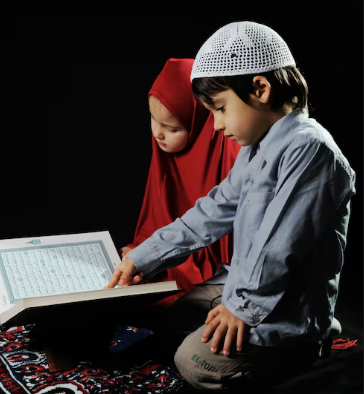Quran Memorization for kids A young boy wearing a white kufi cap and a young girl in a bright red hijab sit together against a black background, focusing intently on an open Quran placed on a wooden stand. The boy points toward the Arabic text as they study.