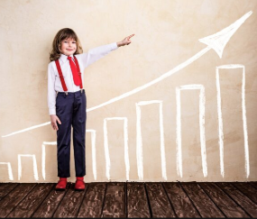 Screenshot 2026-05-16 114304 A young child dressed in business attire—a white shirt, red tie, red suspenders, and dark trousers—stands on a wooden floor against a beige wall. The child smiles and points towards a large, white hand-drawn bar graph with an upward-trending arrow, symbolizing financial or personal growth.