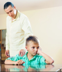Family disadvantages A man with long hair tied back stands behind a young boy who is sitting at a wooden table. The man places a comforting hand on the boy's shoulder, while the boy rests his chin on his fist with a sad, frustrated expression, looking away from the camera.