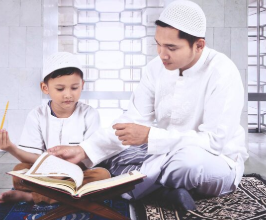 A man and a young boy, both dressed in white thobes and white knit kufi caps, sit cross-legged on prayer rugs. The man gently turns the page of an open Quran resting on a wooden stand in front of the boy, who holds a yellow pencil and watches attentively.