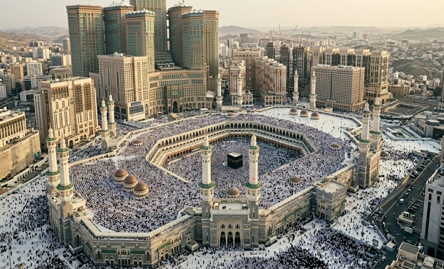 An aerial high-angle view of the Masjid al-Haram in Mecca during daytime, showcasing the holy Kaaba centered within the large white marbled courtyard filled with a vast crowd of pilgrims, surrounded by grand architecture and the Abraj Al Bait towers looming in the background.