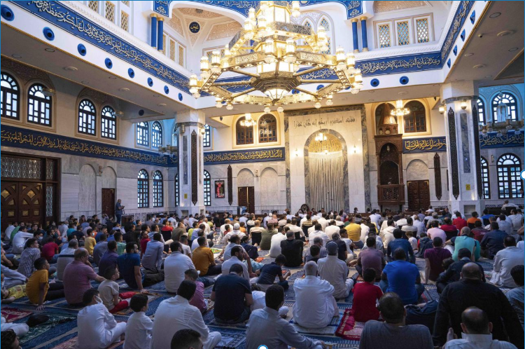 An interior view of a beautifully decorated mosque with blue and white calligraphic walls, capturing a large congregation of men and children sitting on patterned carpets under a massive ornate chandelier during a gathering.