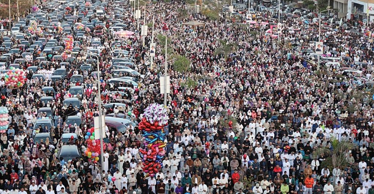 A high-angle, wide aerial photograph capturing a massive, dense crowd of thousands of Muslim worshippers performing Eid prayer outside across a wide multi-lane city street lined with parked cars and colorful balloon vendors.