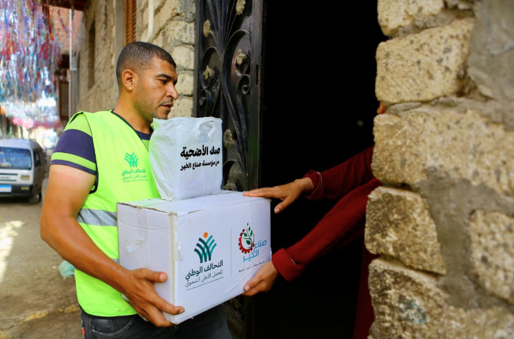 A volunteer man wearing a bright neon green safety vest carries a large cardboard delivery box and a plastic bag marked "Udhiyah Box" in Arabic, handing them over to a resident at a stone doorway.