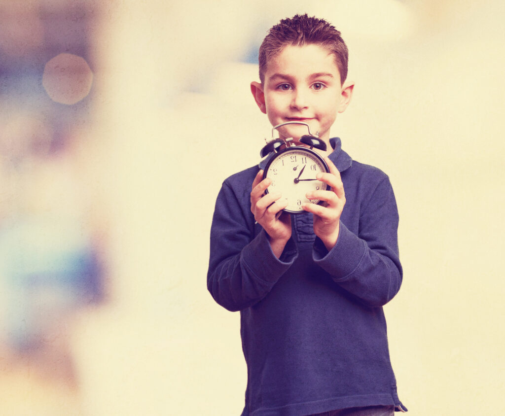 little kid with an alarm-clock A young boy with short hair and a navy blue polo shirt stands smiling while holding a classic black twin-bell alarm clock with both hands in front of his chest, against a softly blurred, light background.