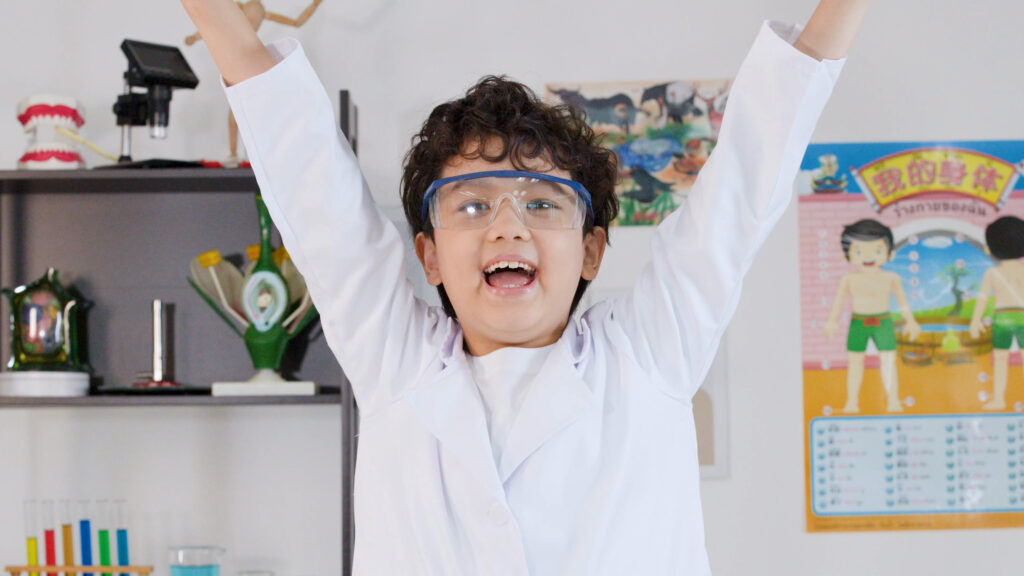 A young East Asian boy with curly hair, wearing a white lab coat and clear safety goggles, stands with his arms triumphantly raised in the air, smiling with an excited, open-mouthed expression inside a science classroom.