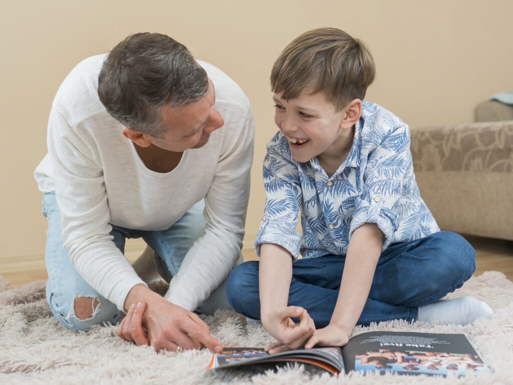 "Learning with Laughter: The Power of Joyful Connection in Early Education" A father in a white long-sleeve shirt and blue jeans leans in to look warmly at his young son, who is sitting cross-legged on a fluffy rug, laughing while reading an open book on the floor.