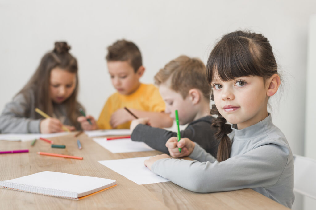 girl-painting-with-friends A young girl with dark bangs and a side braid sits at a wooden table, looking directly at the camera with a gentle smile. In the blurred background, three other children are sitting in a row, focused on drawing on sheets of paper with markers and colored pencils.