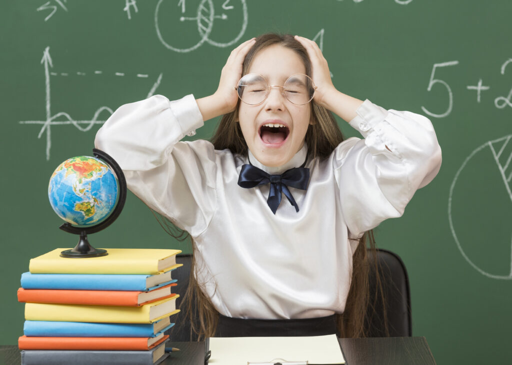 shouting-young-girl-medium-shot A young schoolgirl wearing round glasses sits at a desk, clutching her head and shouting with her eyes squeezed shut in frustration. In front of her is a tall stack of colorful textbooks topped with a mini globe, while a green chalkboard covered in math equations and diagrams serves as the background.
