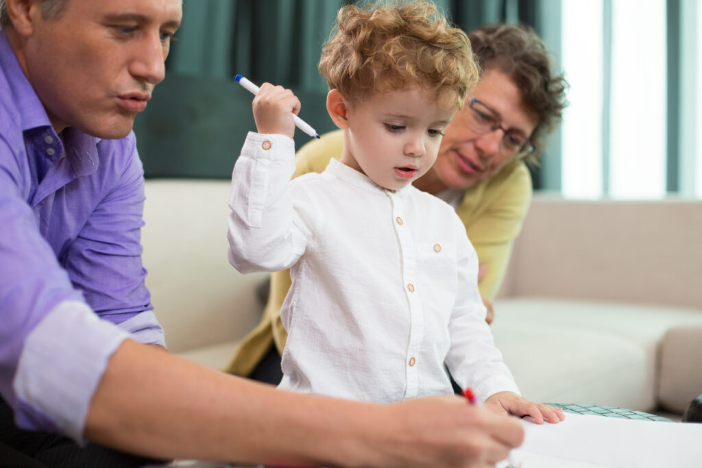Granny, Little Boy and Father Coloring Album A young, curly-haired toddler in a white button-up shirt holds a blue marker raised in the air, looking down curiously at a piece of paper. Beside him, a man in a purple shirt leans forward to demonstrate drawing, while a woman with glasses watches encouragingly from the background in a living room setting.