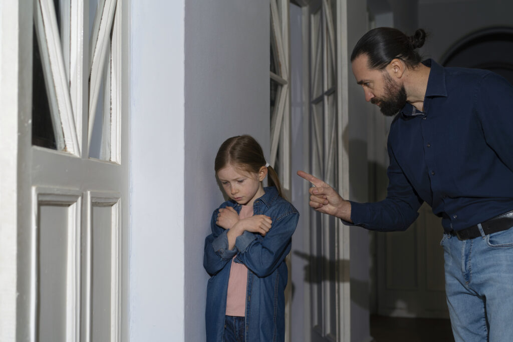young-child-receiving-emotional-abuse-from-parent-home A man with a bun and beard stands over a young girl in a hallway, pointing his finger at her in reprimand. The young girl stands huddled with her back against a white wall pillar, crossing her arms defensively with a sad, downcast expression.