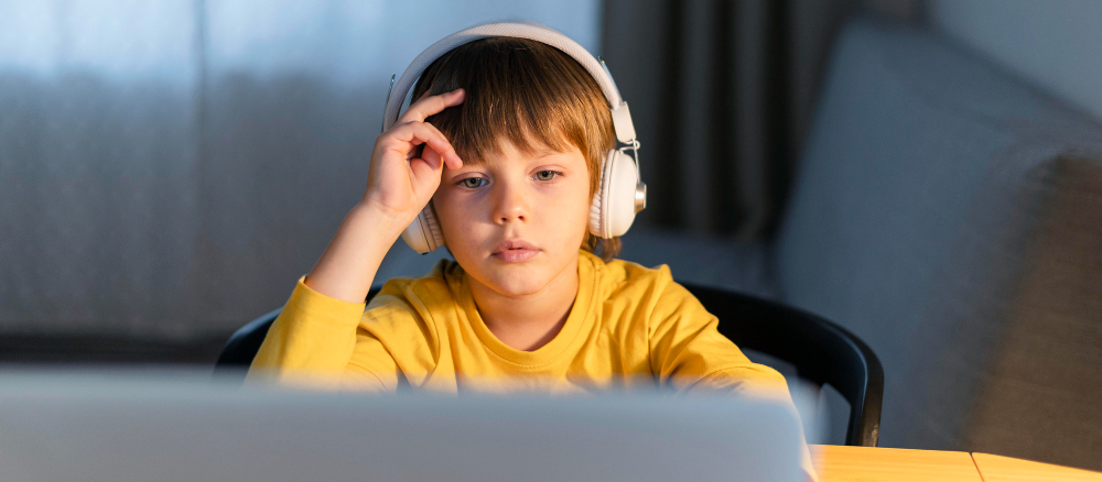 Ages 6–8 A Strong Age to Start Online Quran Classes A professional horizontal close-up photograph of a focused young boy wearing white headphones and a yellow shirt while sitting at a desk and looking at a laptop screen, illustrating why ages 6–8 a strong age to start online Quran classes.