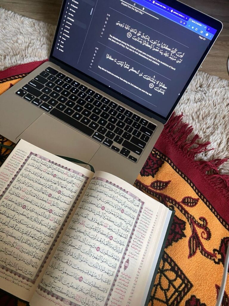 Benefits of Online Tajweed Classes for Kids Top-down view of an open physical Quran with Arabic script resting on a patterned prayer mat next to an open laptop displaying digital Quran verses with English translation.
