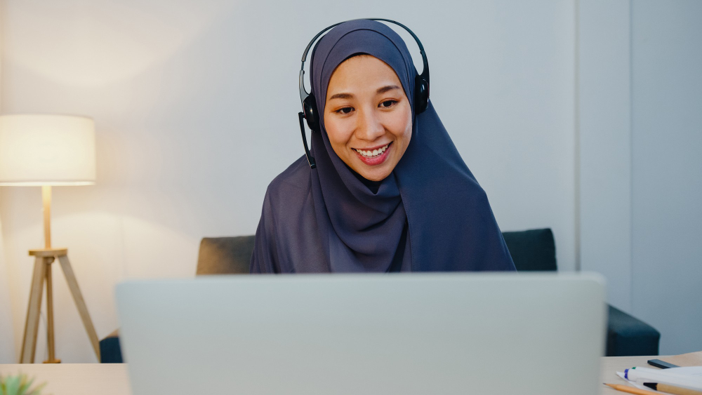 A high-quality lifestyle photograph of a smiling Muslim woman wearing a grey hijab and a headset, looking at her laptop screen while conducting a virtual class, representing the concept of best online quran teachers for kids.