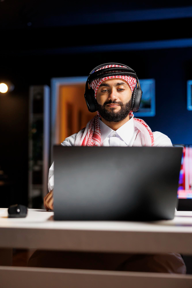 Decide Whether Online Quran Classes Can Actually Work A professional vertical portrait of a Muslim man in a red and white ghutrah and headphones looking at his laptop, concept image for parents trying to decide whether online Quran classes can actually work for their kids.