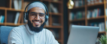 A high-quality panoramic lifestyle photograph of a smiling Muslim male tutor with a beard and glasses, wearing a white thobe, a kufi cap, and a headset while sitting in front of a laptop, capturing the process of how to choose the right private online quran tutor.