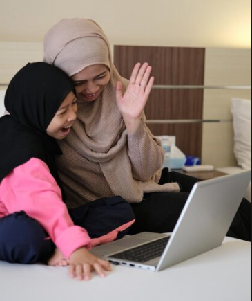 A vertical lifestyle photograph of a smiling Muslim mother wearing a beige hijab waving her hand at a laptop screen while sitting on a bed next to her young daughter who is wearing a black hijab and laughing joyfully during an online session, showcasing online quran academy trust and the parent decision journey.