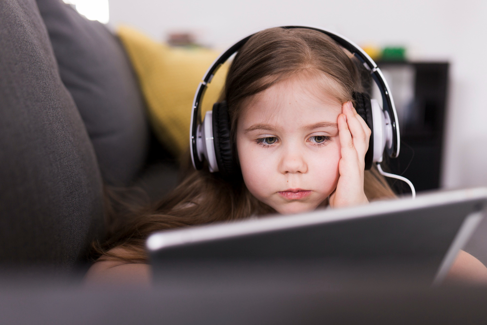 Online Quran Classes Can Help Shy Children Feel More Comfortable A professional close-up photograph of a focused young girl wearing headphones and looking at a digital tablet while resting on a sofa, showing how online Quran classes can help shy children feel more comfortable learning at home.