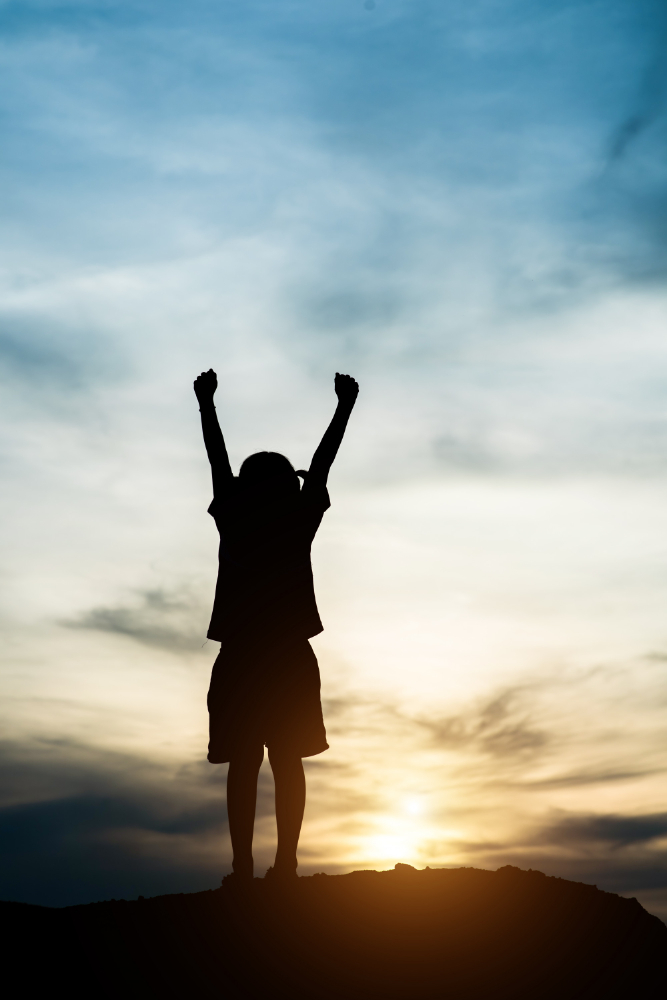 A dramatic vertical silhouette photograph of a young child standing atop a hill with both arms raised high in celebration against a vibrant sunset sky, illustrating the concept of signs an online quran memorization course is working.