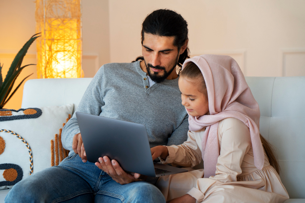 A professional lifestyle photograph of a focused father with dark hair and a beard holding a laptop on a white sofa alongside his young daughter in a pink hijab, illustrating how the first online quran class is part of the bigger parent decision.