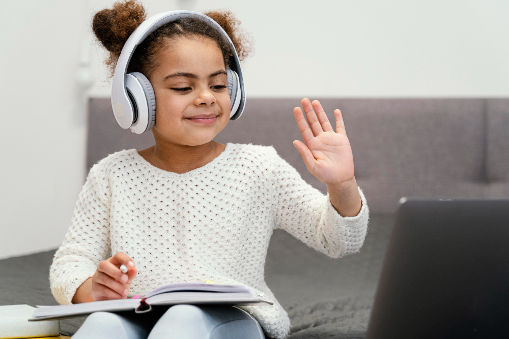 A high-quality lifestyle photograph of a smiling young girl with afro-textured hair buns wearing white headphones and waving at her laptop during a live session, a concept image for what makes the best online quran classes for kids different.