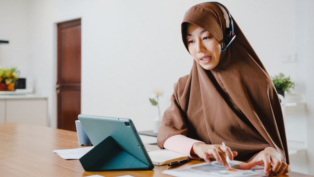 What Progress Looks Like in Online Quran Learning A professional lifestyle photograph of a focused female teacher in a brown hijab and headset using a tablet and analyzing student data sheets, representing what progress looks like in online Quran learning.