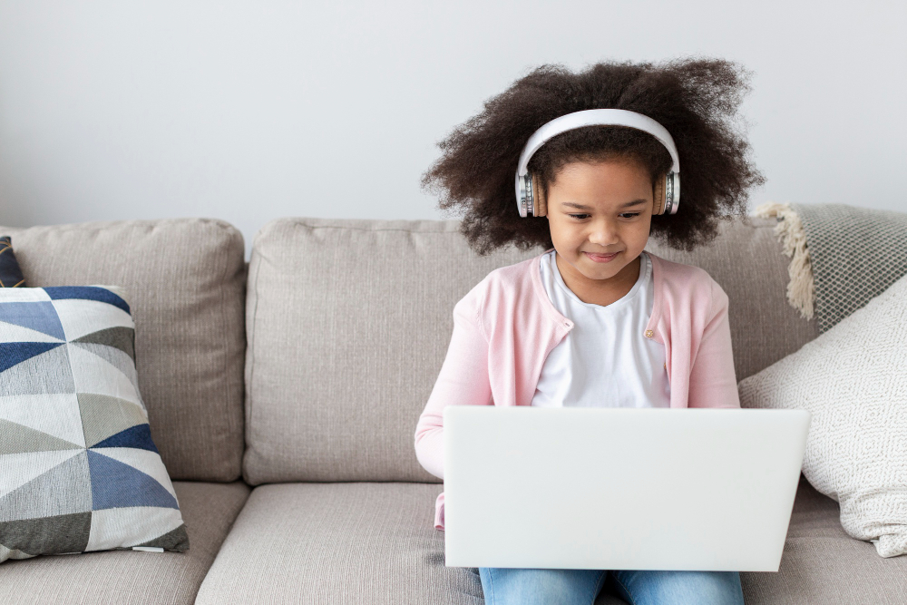 A candid lifestyle photograph of a young Muslim girl with a bright smile wearing a dusty pink hijab, looking directly forward while typing on an open silver laptop at a wooden desk in a bright room, showcasing why Canadian families choose online quran classes for kids.