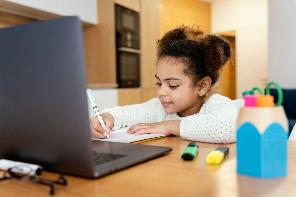 A candid lifestyle photograph of a young mixed-race girl with her hair tied in two high buns, wearing a white textured sweater, sitting at a wooden desk and writing in a notebook with a pen while looking at an open laptop, representing why UK families choose online quran classes for kids.