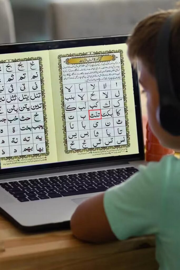 Can You Learn Quran Online Without Knowing Arabic An over-the-shoulder horizontal photograph of a young boy wearing black headphones, sitting at a wooden desk and studying introductory Arabic letters displayed on his laptop screen from a digital Qaida book.