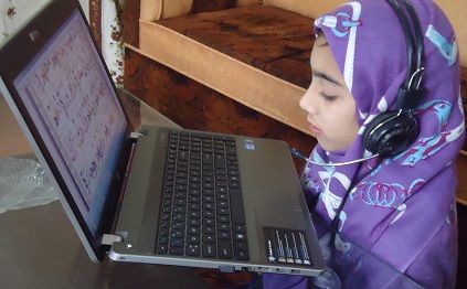 A horizontal lifestyle photograph of a young Muslim girl wearing a purple patterned hijab and a black headset with a microphone, sitting at a desk and looking closely at Arabic text displayed on a laptop screen, demonstrating how can a complete beginner learn quran online.