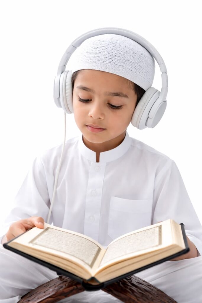 How Do You Learn Quran Online Step by Step A vertical studio photograph of a young Muslim boy with a light complexion wearing a white thobe, a white knit kufi cap, and white headphones, sitting cross-legged while holding and reading from an open Quran placed on a wooden stand.