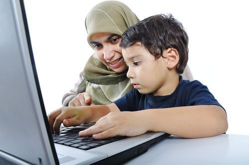 A horizontal studio photograph of a young boy typing on a laptop keyboard while a smiling woman wearing a light green hijab sits behind him and points at the screen, showcasing how long does it take children to learn quran online.