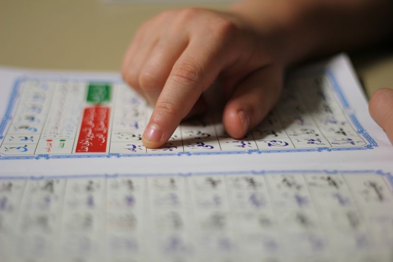 A close-up horizontal photograph of a child's hand with their index finger pointing to Arabic characters in an open Noorani Qaida book, highlighting the focused learning process of Arabic phonics and pronunciation.