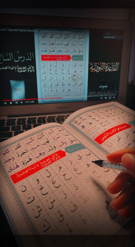 How Noorani Qaida Helps Students Without Arabic A close-up vertical lifestyle photograph of a student's hand holding a pen and pointing at lessons in an open printed Noorani Qaida book, placed in front of an open laptop screen displaying a corresponding video lesson of the same Arabic phonetic tables.