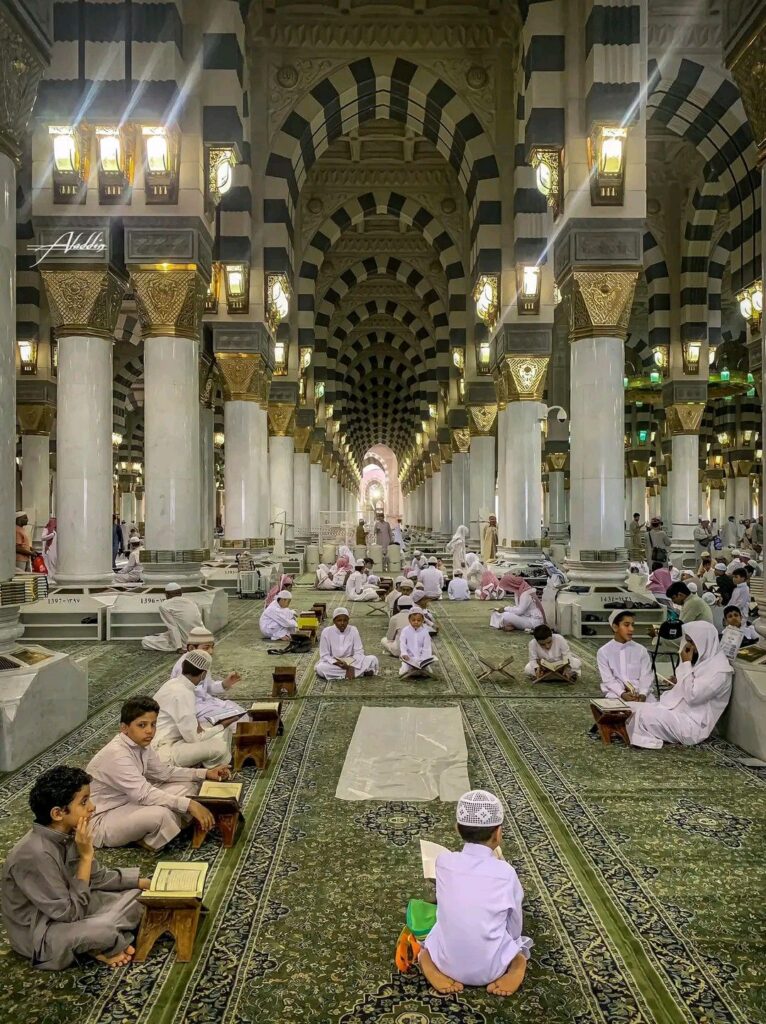 A wide view inside Al-Masjid an-Nabawi in Medina showing rows of children and adults sitting on the green patterned carpet in traditional Islamic attire, reading open Qurans on wooden stands under ornate arches and columns.