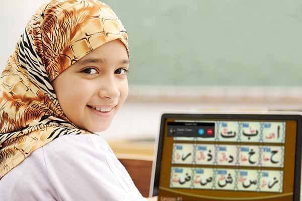 A horizontal lifestyle photograph of a smiling young boy wearing a gold and black patterned head covering, sitting in front of a laptop screen displaying colorful Arabic alphabet cards during an online class.