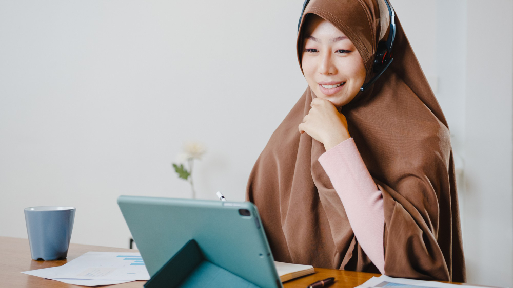 What Makes a Good Online Quran Teacher A smiling female Muslim teacher wearing a brown hijab and a headset with a microphone sits at a wooden desk, looking at a tablet screen while holding a stylus during an online lesson.