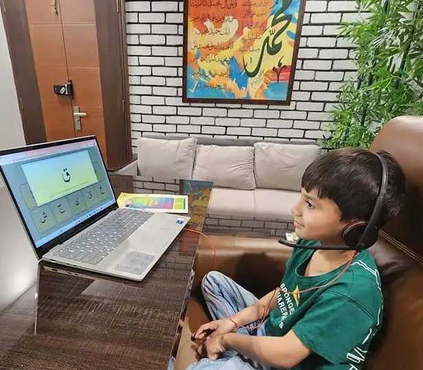 When Should Kids Start Tajweed A horizontal lifestyle photograph of a young boy wearing a green t-shirt and headphones, sitting in a brown armchair and looking at a laptop displaying an Arabic phonics lesson with the letter "Qaf" (ق) on the screen.