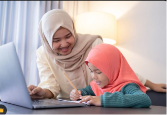 A smiling mother or tutor wearing a beige hijab guiding a young Muslim girl wearing a bright coral pink hijab as she writes notes in front of a laptop computer.