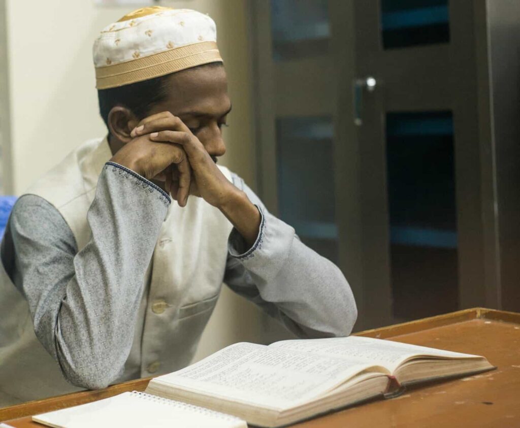 A Muslim man wearing a embroidered prayer cap and traditional vest sitting at a wooden desk with his hands rested under his chin, thoughtfully reading an open book.